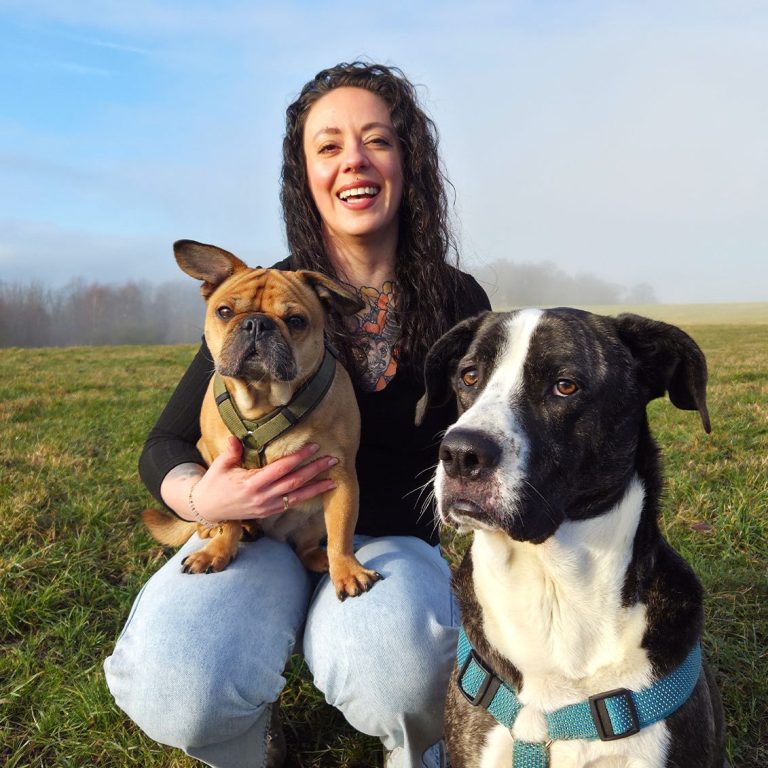 Frau mit lockigem Haar und Tattoo im Dekolletee hockt im Gras und hält einen Hund, neben ihr ein weiterer Hund.