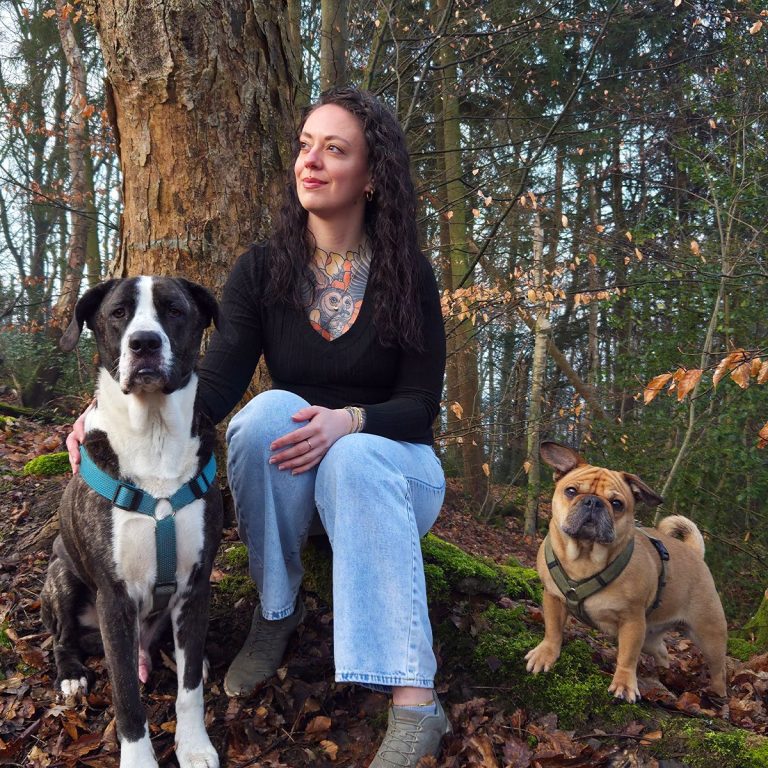 Frau mit Tattoos sitzt im Wald auf einem Baumstamm, neben ihr stehen zwei Hunde, ein großer und ein kleiner.