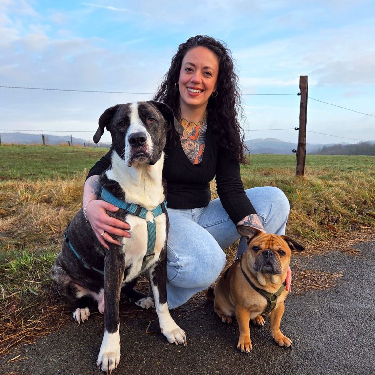 Eine Frau mit lockigen Haaren hockt mit zwei Hunden auf einer Straße, mit Bergen im Hintergrund.